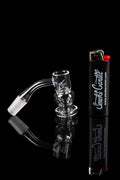 Clear glass dab rig with a quartz banger and lighter, reflected on a black surface with branding.