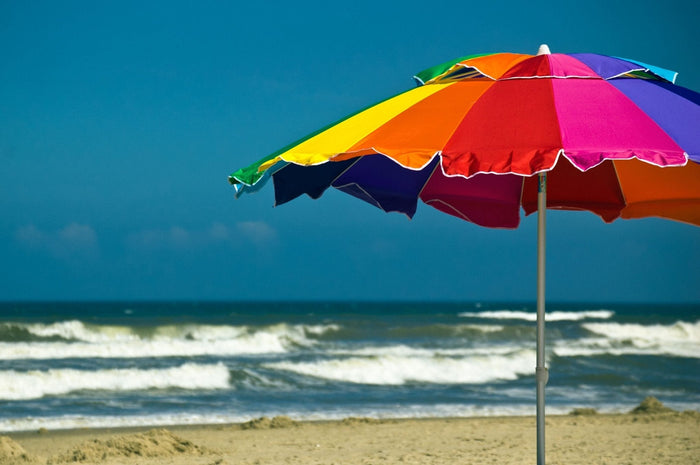 Rainbow 240cm beach umbrella with vibrant rainbow panels, UPF50+ UV protection, and tilt feature on sandy beach near ocean waves