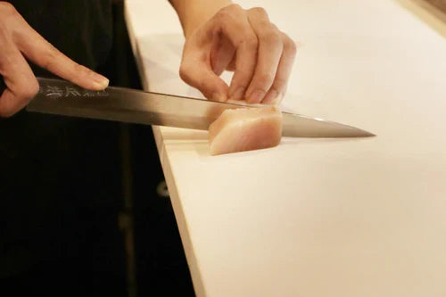 sushi being cut by chefs knife on beige cutting board.