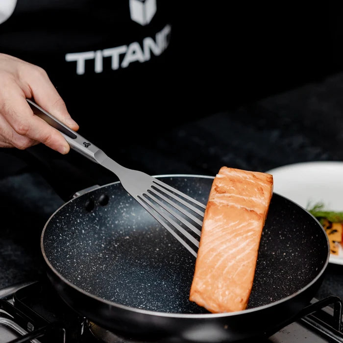 Person cooking salmon in a frying pan with a titanium spatula, wearing a Titanion branded shirt.