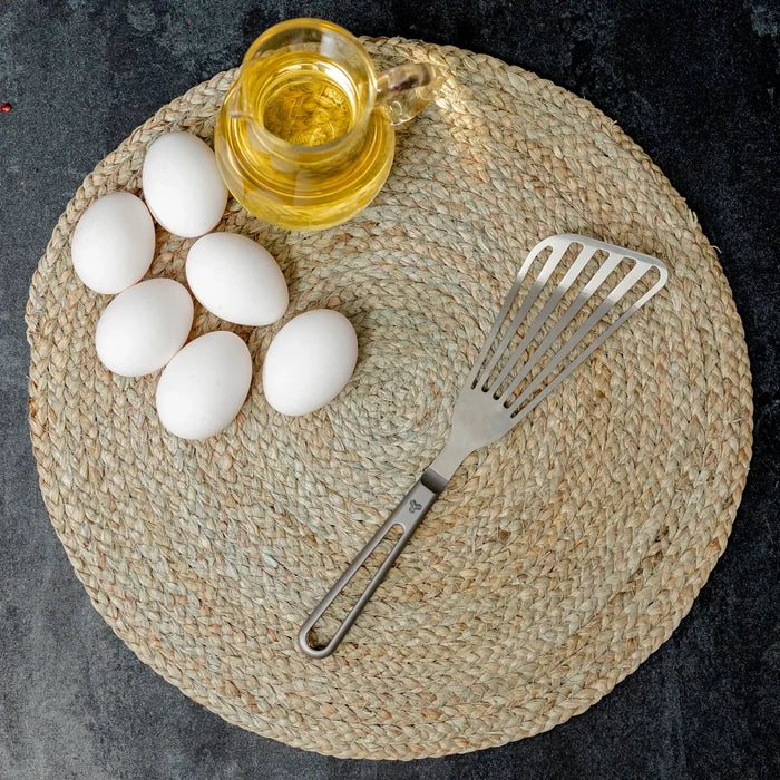 Eggs, oil, and a titanium spatula on a woven mat.